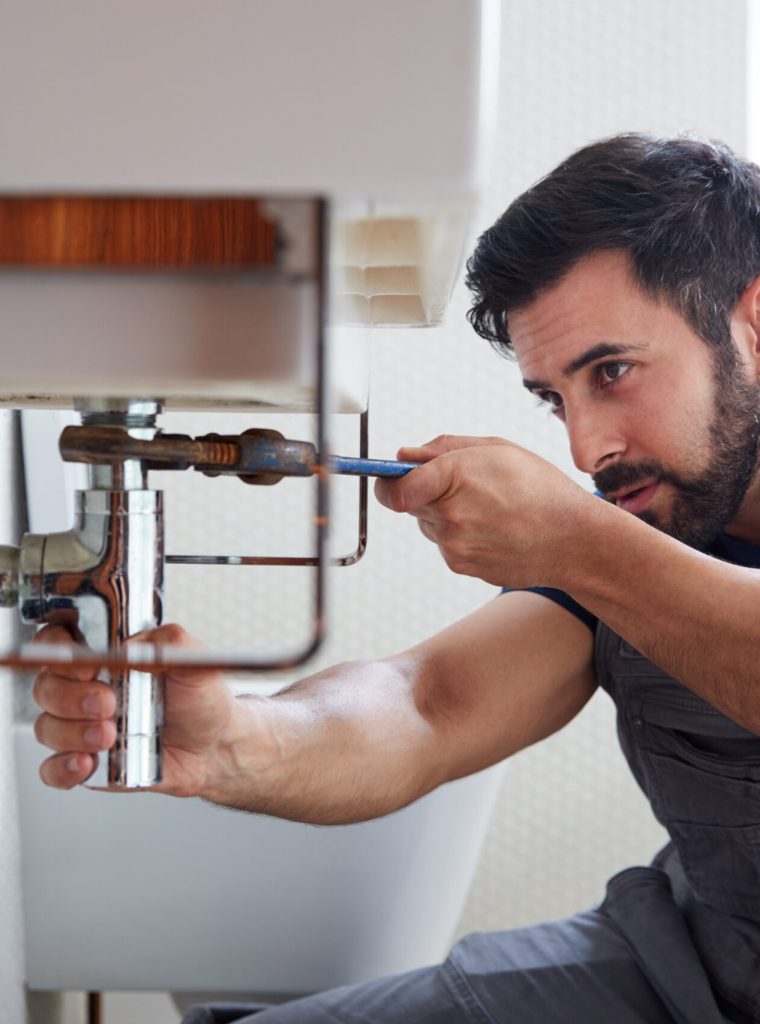 Male Plumber Using Wrench To Fix Leaking Sink In Home Bathroom
